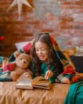 Child reading a book under a plaid blanket with a teddy bear, next to a decorated Christmas tree against a brick wall.