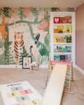 Playroom with painted mural with kids names, tiger, toucan, sloth, monkey. Bookshelf with books arranged by color. kids play kitchen and wood slide