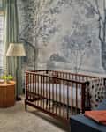 Nursery with a wooden crib, beige armchair, green curtains, and nature-themed wallpaper featuring trees.