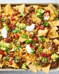 overhead shot of nachos in a sheet pan,  topped with cilantro, avocado, sour cream and salsa.