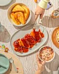 A table set with a meal featuring barbecue chicken, macaroni and cheese, coleslaw, and cornbread, with drinks and utensils.