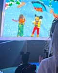 A child with a white bow in their hair watches a colorful stage performance featuring Mickey Mouse and friends.