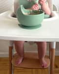 A baby in a high chair playing with a green bowl of food, with bare feet visible on the wooden footrest.