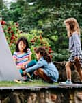 Three children playing near a white cardboard playhouse in a garden with red flowers.