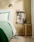 Cozy bedroom corner featuring a green and white polka dot bedding set, a wall-mounted organizer with sleep mask, notes, and a water bottle, alongside a wooden side table with a glass and book.