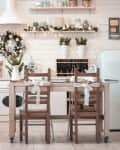 Cozy kitchen with white cabinets, wooden table, festive wreaths, and string lights, featuring a vintage fridge and decor.