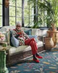 Elderly woman in colorful attire sitting on a wicker sofa in a sunroom with large plants and vibrant decor.