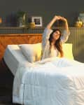 Woman stretching in bed with white bedding, wooden headboard, and a plant by a sunlit window.