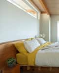 Minimalist bedroom with wooden beams, a bed with yellow sheets, white quilt, and a wooden bench with folded towels and books.
