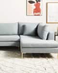 Light blue sectional sofa with gold legs on a textured rug, framed art, and a small side table with a lamp.