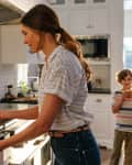 Woman removing grilled cheese from oven in a bright kitchen, child eating in the background.
