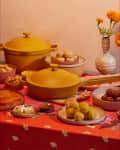 Yellow cookware set on a red patterned tablecloth with assorted Indian sweets and marigold flowers in a striped vase.