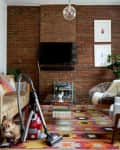 Living room with brick wall, tan sofa, colorful rug, vacuum, and a small dog. Wall art and plants add decor.