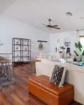 Cozy living room with white sofas, wooden floors, ceiling fans, plants, and a large window with natural light.