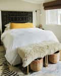 Bedroom with a black patterned headboard, white bedding, yellow pillow, fur bench, woven baskets, and wall art.