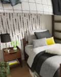 Dorm room with a loft bed, patterned bedding, nightstand with lamp, books, fan, and cactus on the windowsill.