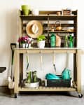 Gardening station with tools, potted plants, a hat, and a bench with striped cushions against a white wall.