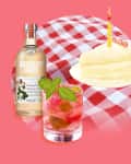 Absolut Juice bottle, strawberry cocktail with mint, and a slice of cake with a candle on a checkered cloth.