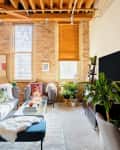 Loft living room with exposed brick, large windows, modern furniture, plants, and a mix of colorful and neutral decor.