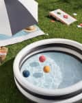 Inflatable pool with colorful balls on grass, next to a striped blanket, sun hat, and cornhole game.