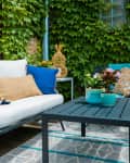 Outdoor patio with white cushioned sofa, blue and striped pillows, black table, potted plants, and ivy-covered brick wall.
