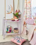 Lilac cart with books, plant, and decor beside floral curtains and a crescent moon artwork in a cozy room corner.
