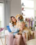 Woman in floral dress sitting on pink chair, hugging a golden retriever with a blue bow tie, in a bright room.