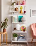 White ladder shelf with books, globe, plants, and decor items next to a pink chair and framed geometric art.