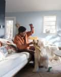 Woman playing with a dog in a bright living room with a beige sofa, round wooden table, and large potted plant.