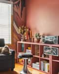 Cozy living room with a black sofa, colorful pillows, a dog, and a pink shelf with books, plants, and a stereo.