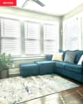 Sunroom with blue sectional sofa, patterned rug, potted plant, and ceiling fan, featuring large windows with blinds.