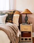Bedroom with a beige bed, floral pillows, a woven blanket, and a wicker nightstand with a lamp.
