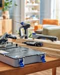 A woodworking setup featuring a circular saw, rotary tool, and drill on a table with stacked plywood sheets in a bright room.