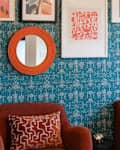 Colorful living room corner featuring teal patterned wallpaper, orange round mirror, and two red armchairs with decorative pillows.