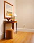 A wooden console table with a framed landscape painting, a lamp, a bowl of apples, and a round orange stool on hardwood flooring.
