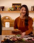 A woman in an orange top serves a cake in a modern kitchen with a charcuterie board, fruits, and decorative items on shelves.