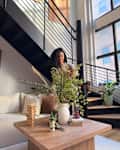 A woman with curly hair arranging a floral centerpiece on a wooden coffee table in a modern living room with large windows.