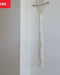 Macramé wall hanging with a wooden rod, displayed on a light-colored wall.