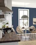Cozy kitchen dining area with a round white table, rattan chairs, and a dark blue wall, featuring a pendant light and greenery.
