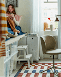 Cozy bedroom corner with a woman sitting on a bed, holding a mug, surrounded by a desk, chair, and colorful rug.