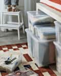 Clear storage bins filled with folded clothes under a bed, with a white step stool and a patterned rug nearby.