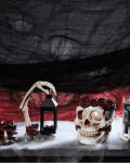 Skull candle holder with rose crown, skeleton hand lantern, and black candle on smoky surface with red and black backdrop.