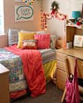 Cozy bedroom with a colorful quilted bedspread, decorative pillows, a desk with books, and a camera hanging on the wall.