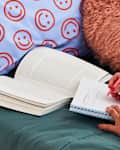closeup shot of girl laying on a bed with teal sheets, smiley face pillowcase  and a furry circled pillow writing in a notebook with a pink pen and a text book in front of her