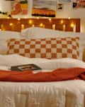 Cozy bedroom with a wooden bed, checkered pillow, orange throw blanket, and decorative plants on a nightstand.