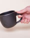 Hand holding a dark ceramic mug with a speckled finish.