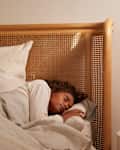 Person sleeping in bed with a wooden headboard, next to a glowing alarm clock on a bedside table.
