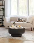 Beige sofa with patterned cushions, round black coffee table, and floor lamp in a bright living room with large windows.