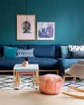 Modern living room with teal wall, blue sectional sofa, patterned cushions, pink ottoman, and a beige rocking chair.