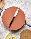 Terracotta frying pan with lid on terrazzo surface, surrounded by plates, utensils, garlic, shallots, and fresh vegetables.
